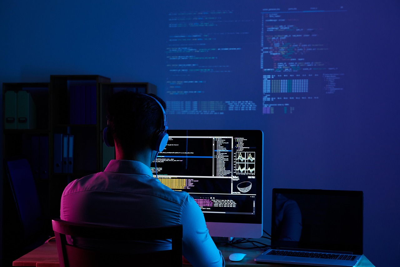 A developer wearing headphones working at a desk with a monitor and laptop showing code in a dark blue room